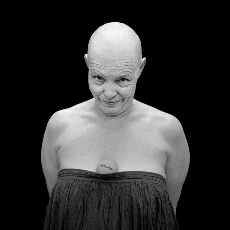 Black-and-white portrait of a bald person in a strapless dark top, smirking against a black background
