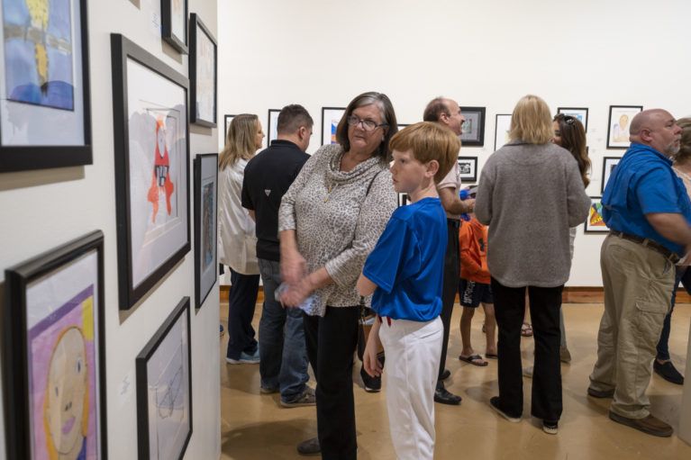 People browsing framed artwork in a bright gallery, with several standing and chatting near the walls.