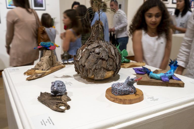 Small sculptural artworks displayed on a table at an indoor exhibition, with people browsing in the background