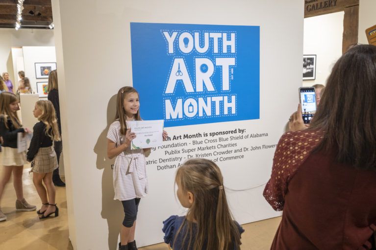 Youth Art Month exhibit with a blue sign, children viewing displays in a gallery