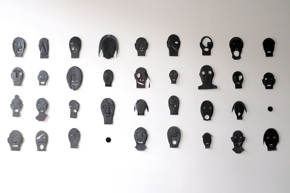 Rows of small black and gray face-like masks or figurines arranged on a white wall.