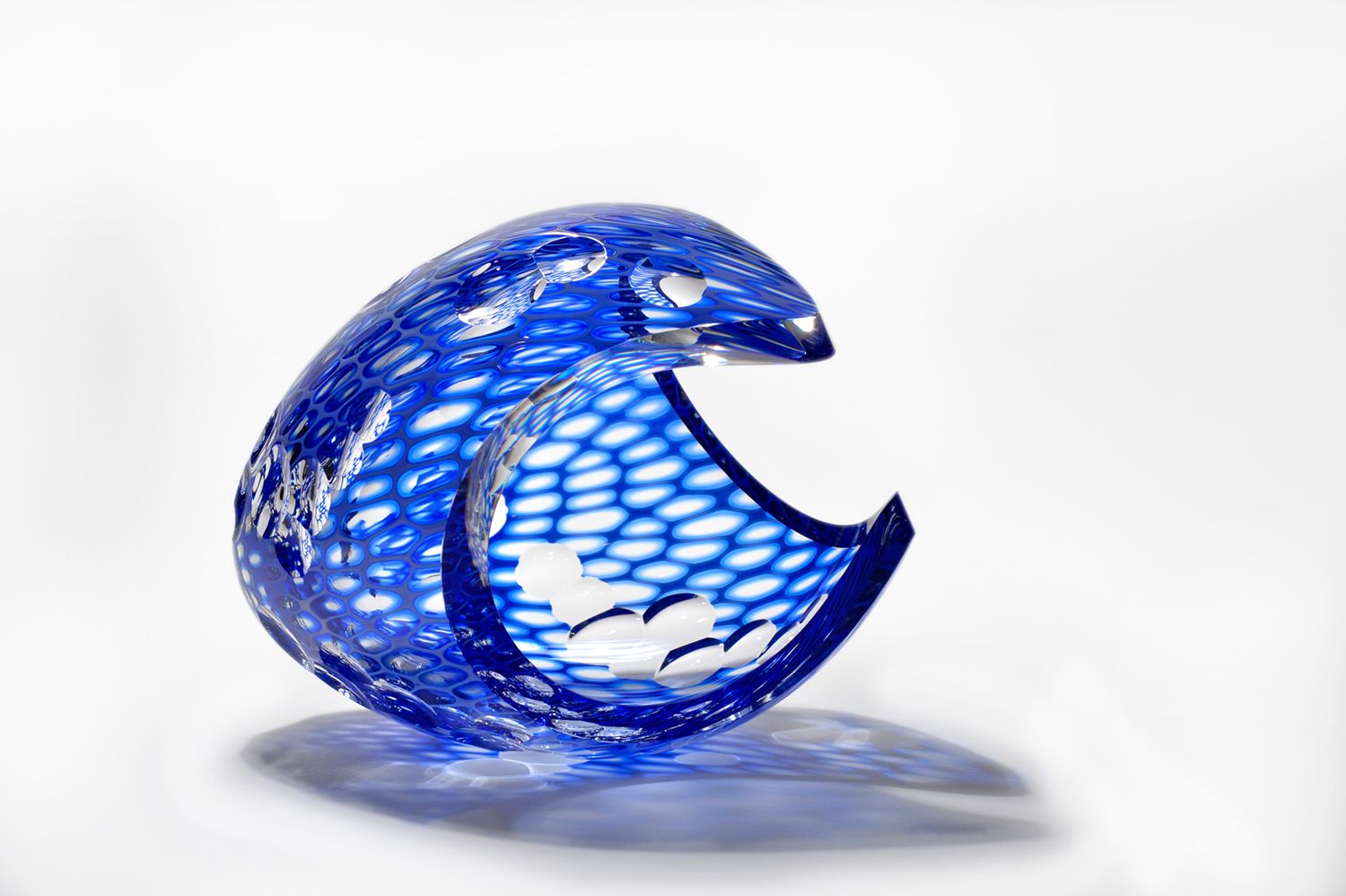 Blue glass sculpture shaped like a hollow crescent with a textured, net-like surface on a white background