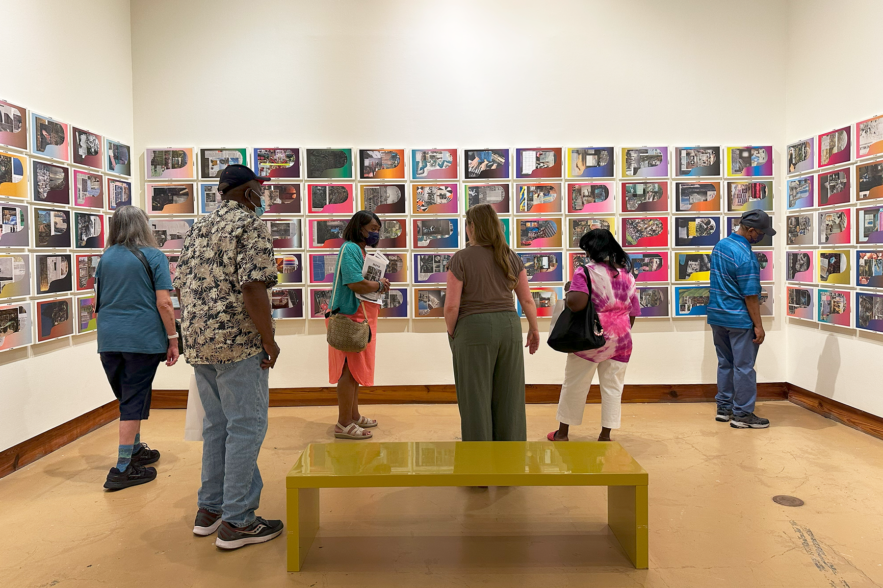 People viewing colorful wall art in a gallery with a yellow bench in the foreground