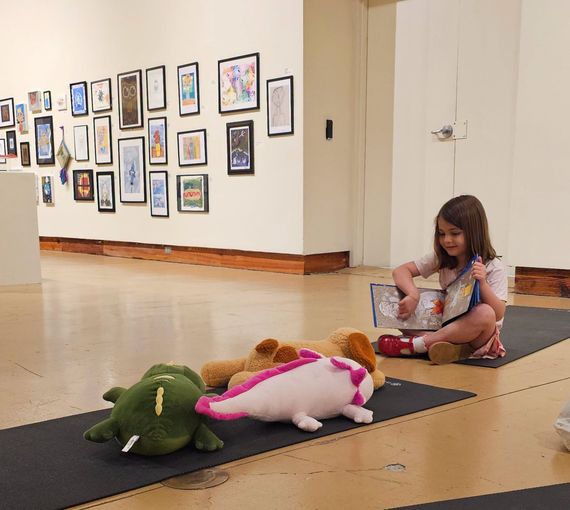 Child reading on the floor beside plush toys in an art gallery hallway with framed artwork on the wall