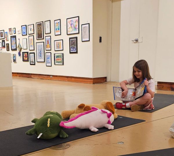 Child reading on the floor beside plush toys in an art gallery hallway with framed artwork on the wall