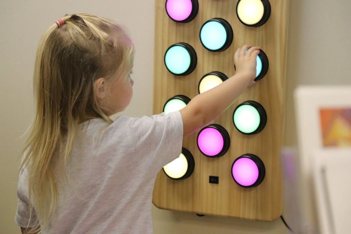 Child reaching for a lit touch panel with colored circular buttons on a wall