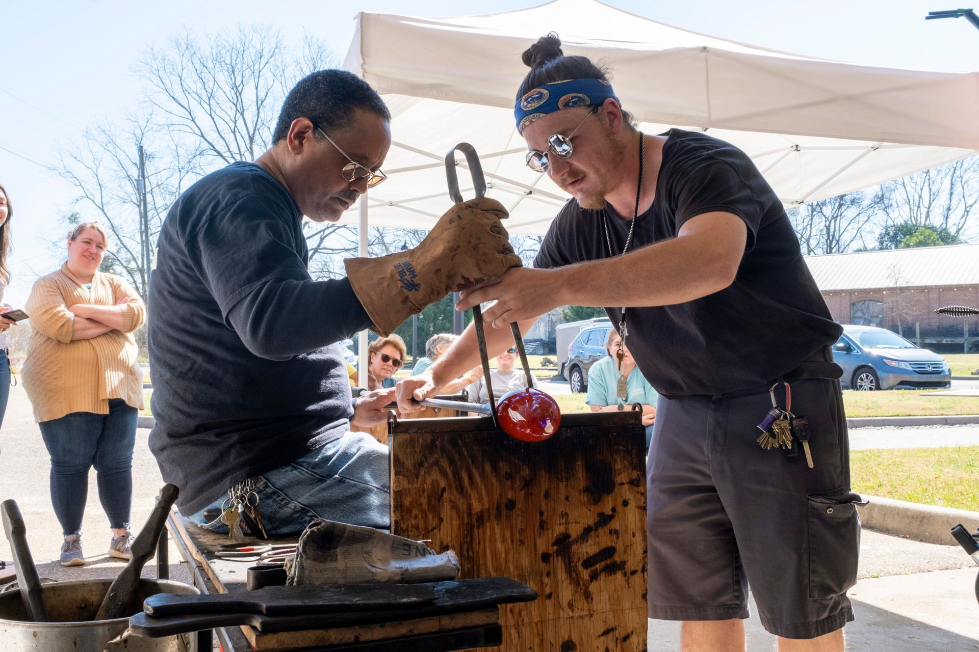 Two glassblowers shaping molten glass on a workbench under a canopy outdoors.