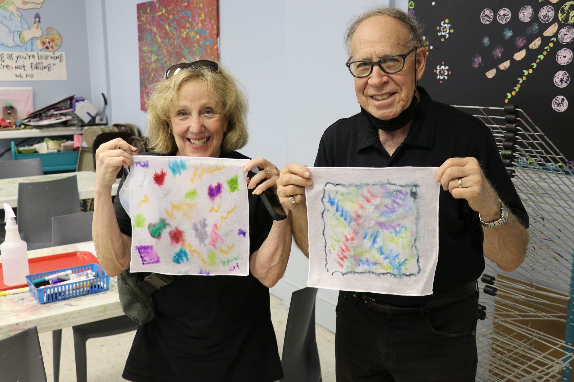 Two people holding colorful abstract art pieces in an art studio, smiling at the camera.