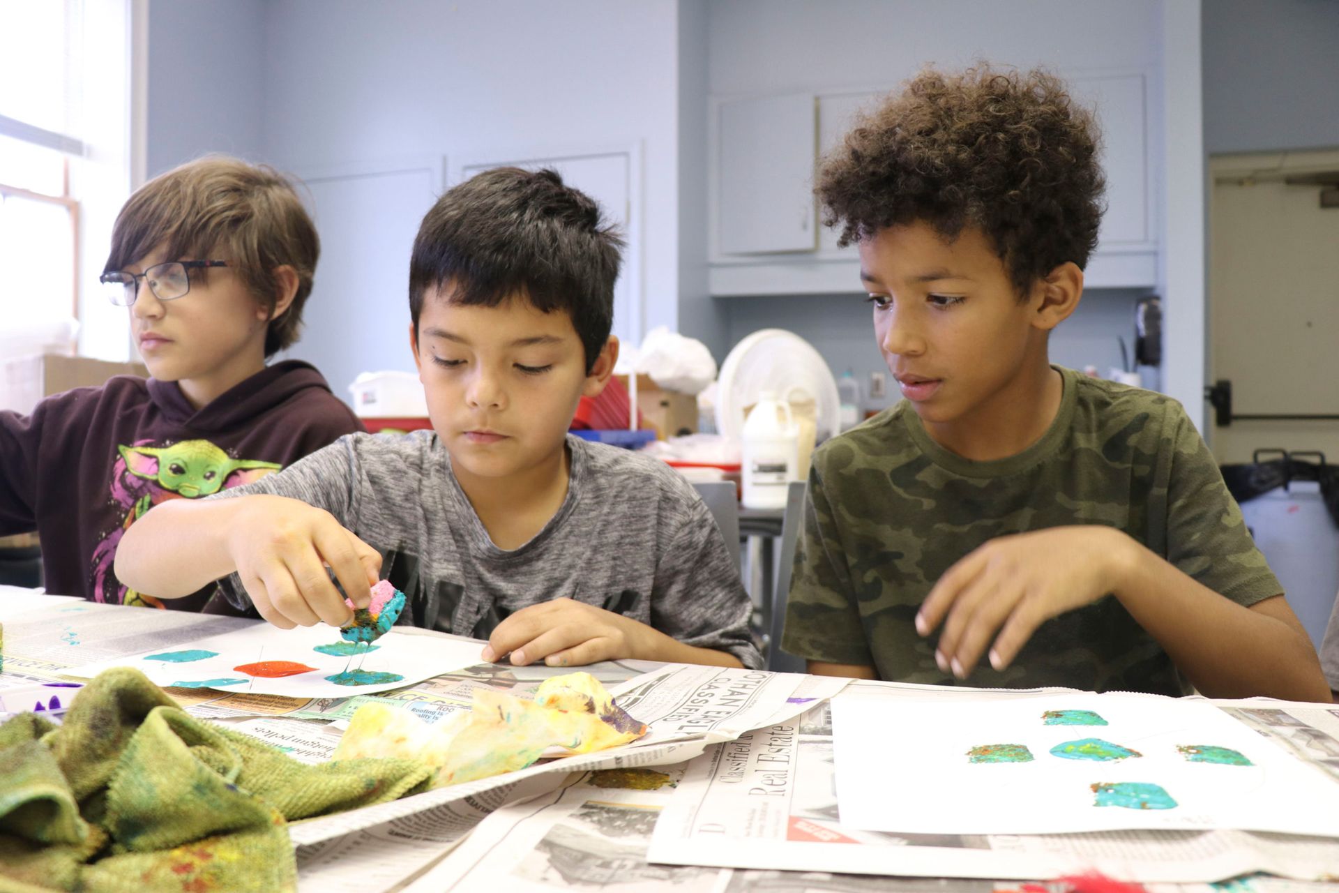 Three children seated at a table, painting and working on art projects in a classroom.