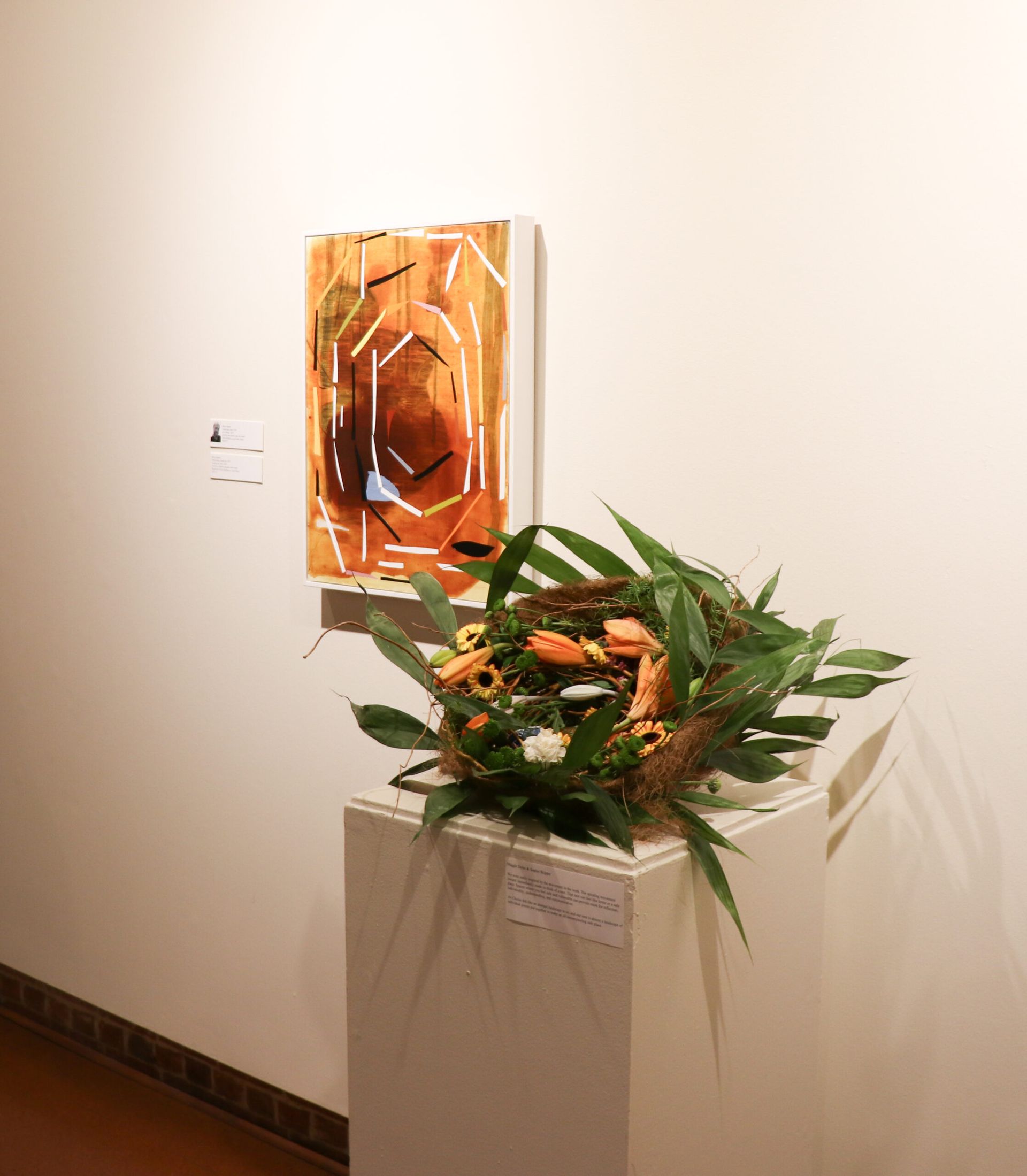 Wall-mounted orange artwork above a white pedestal with a green floral arrangement in a gallery space