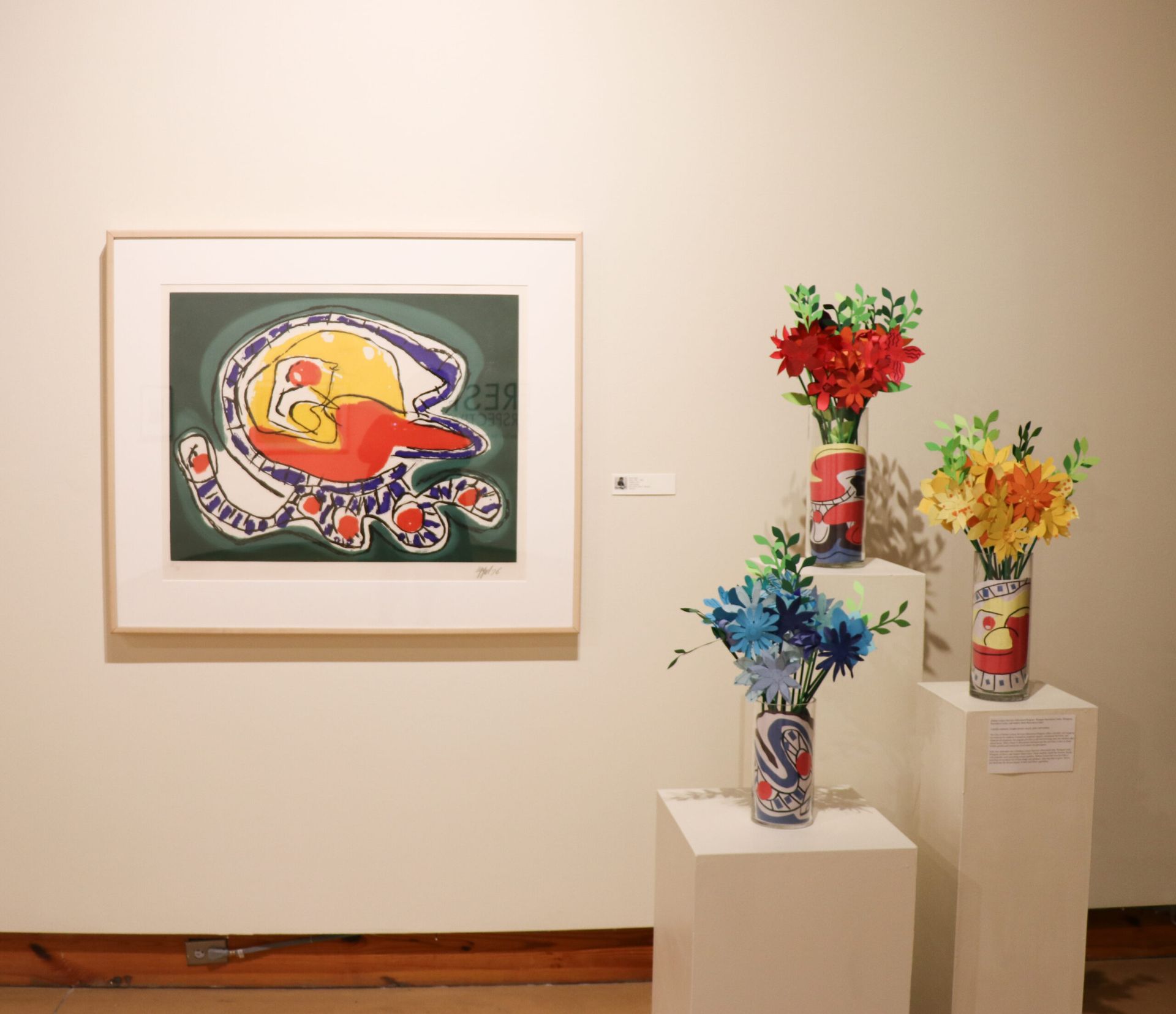 Art gallery wall with a framed colorful painting and three vase sculptures on pedestals.