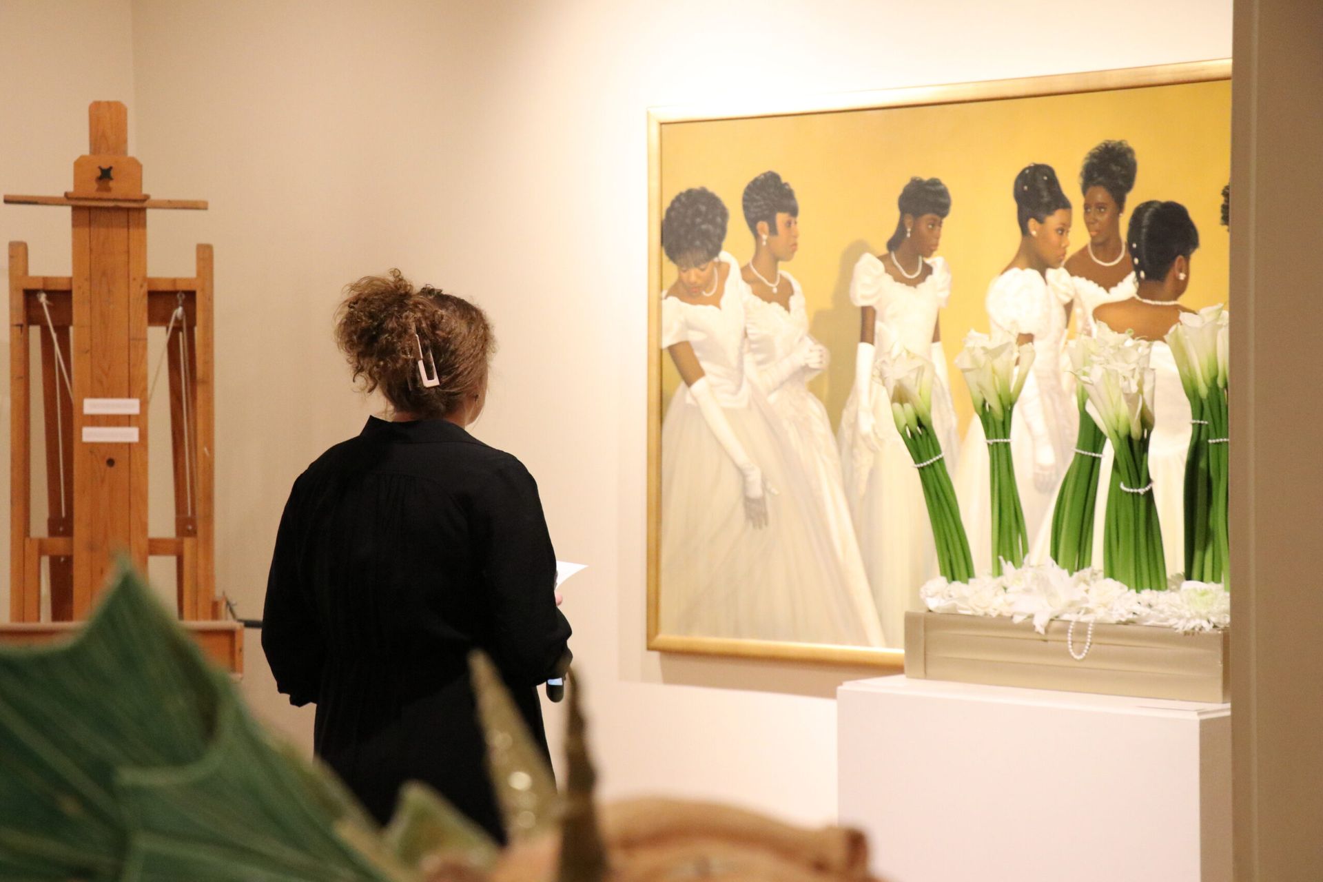 Person viewing a gallery painting of women in white dresses beside a wooden sculpture