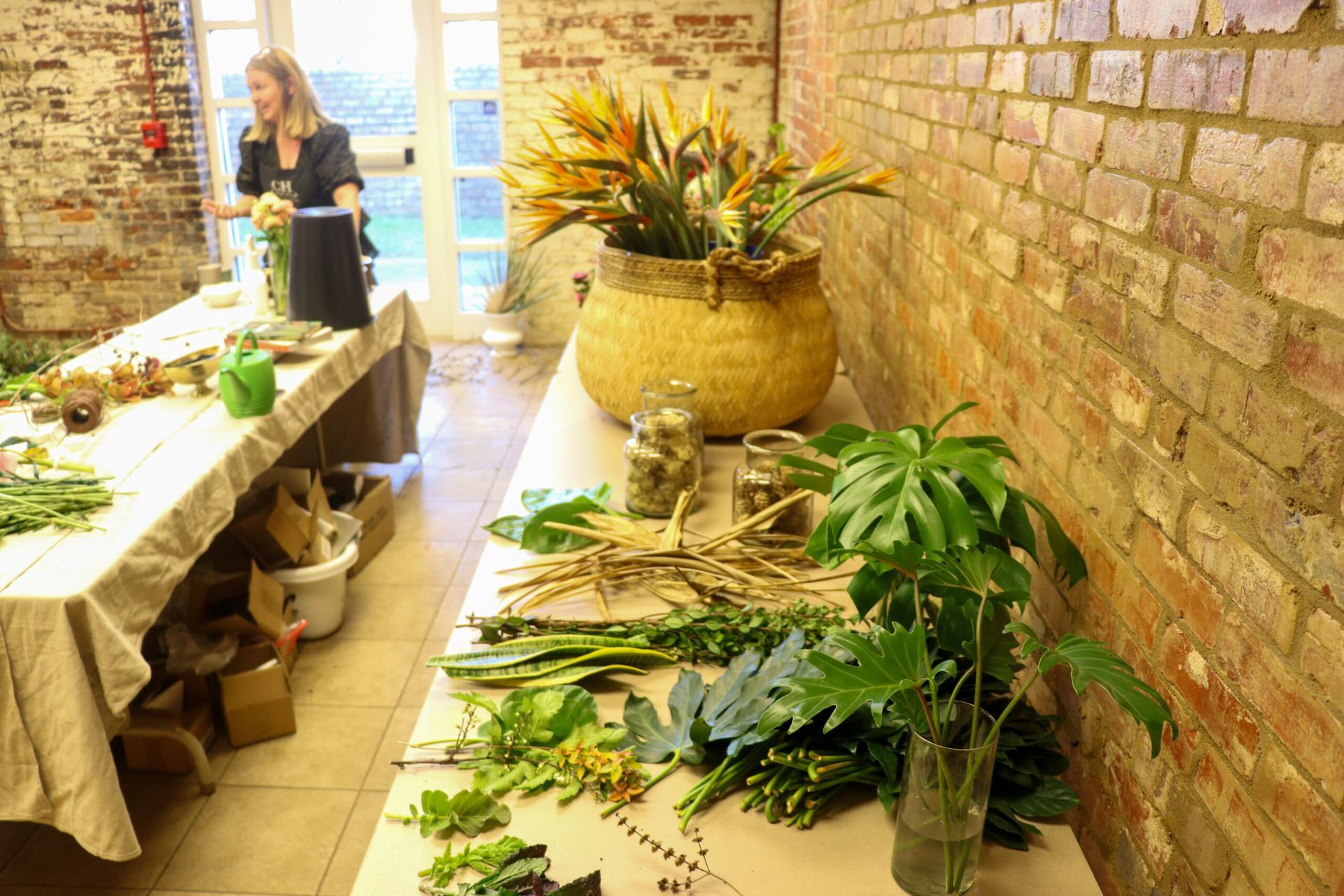 Woman arranging flowers in a rustic stone room with long tables, pots, and plants