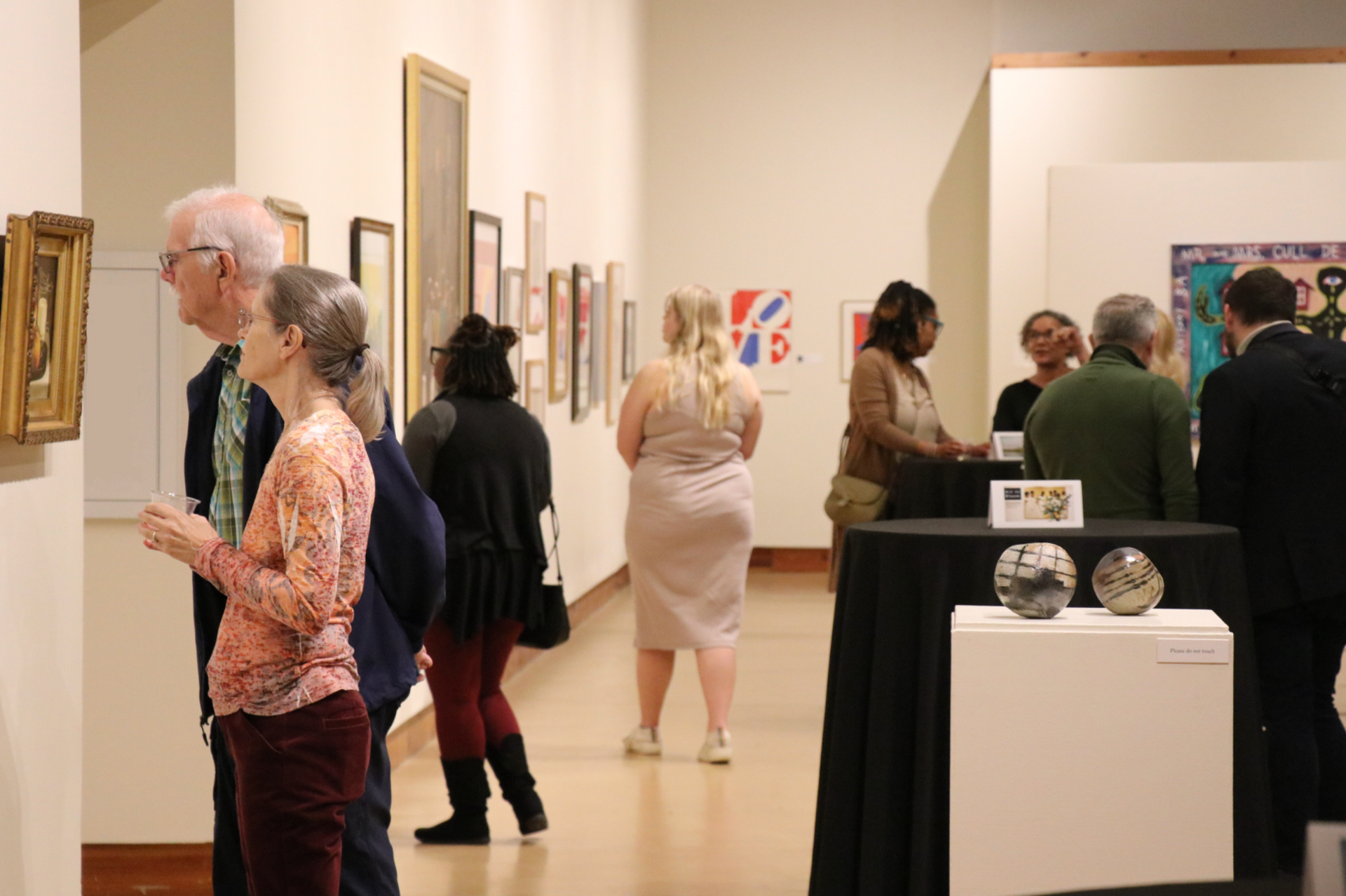 People viewing art in a gallery, with framed paintings on the walls and sculptures on display.