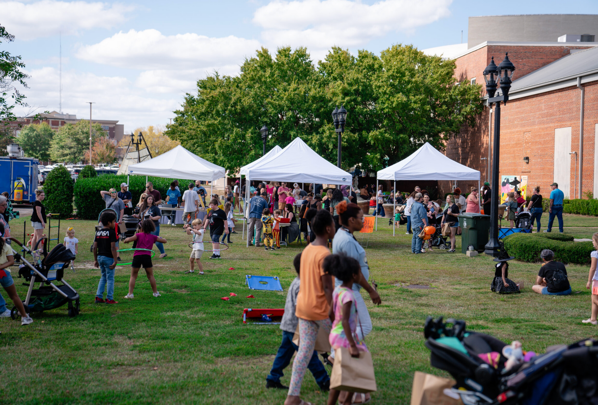 People mingling on a grassy lawn at an outdoor community event with white tents and a brick building.