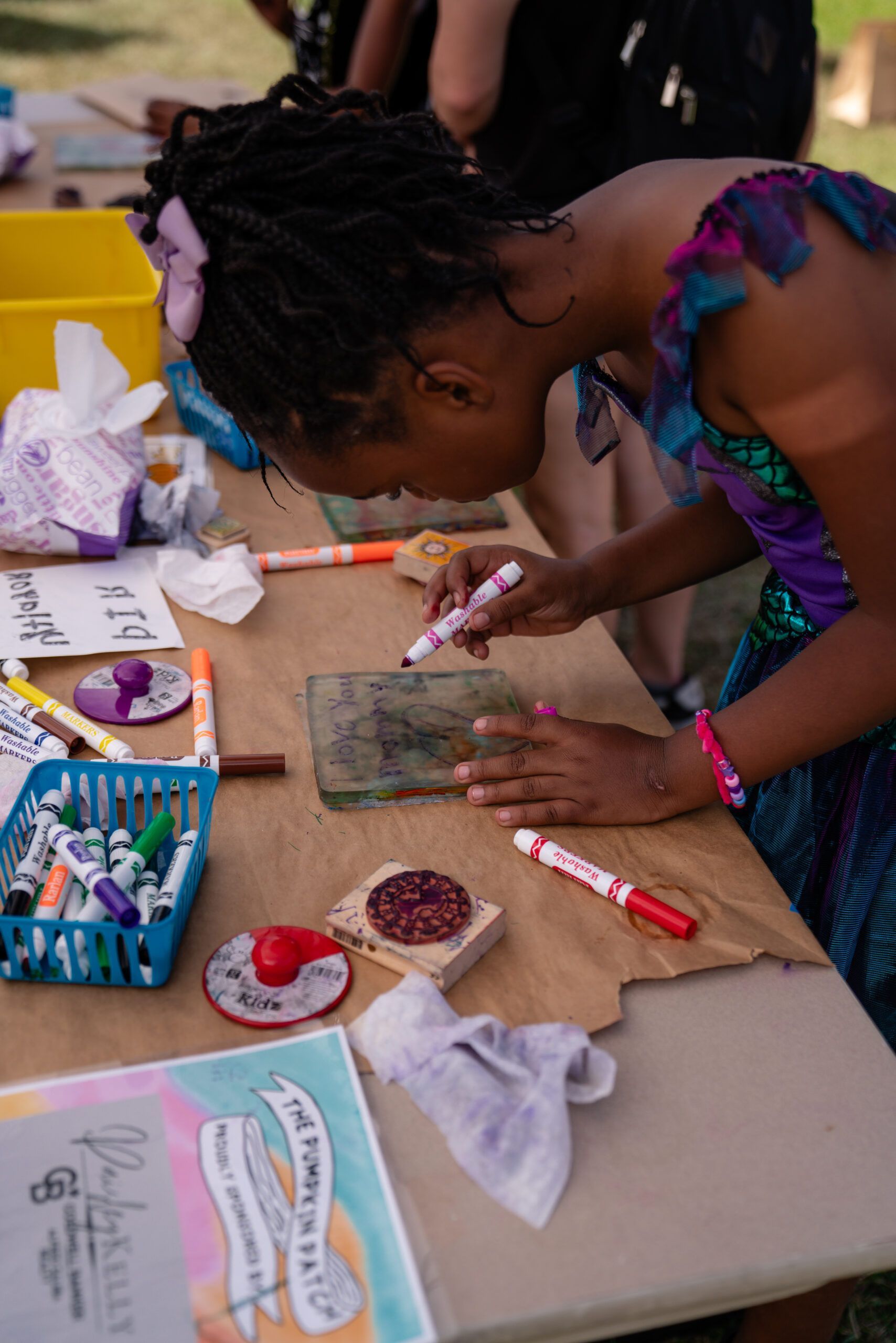 Person painting a small tile at an outdoor craft table with markers and paint supplies nearby