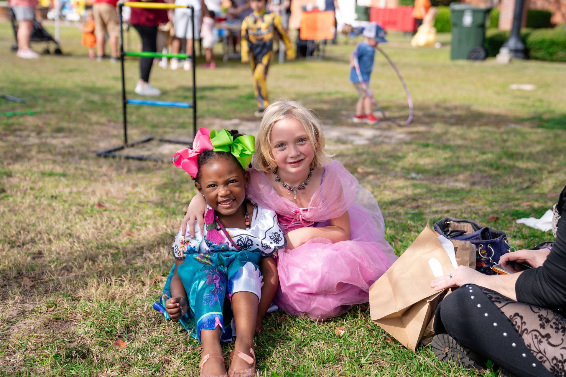Two children smiling on grass at an outdoor event, one in a pink tutu and the other in a blue dress