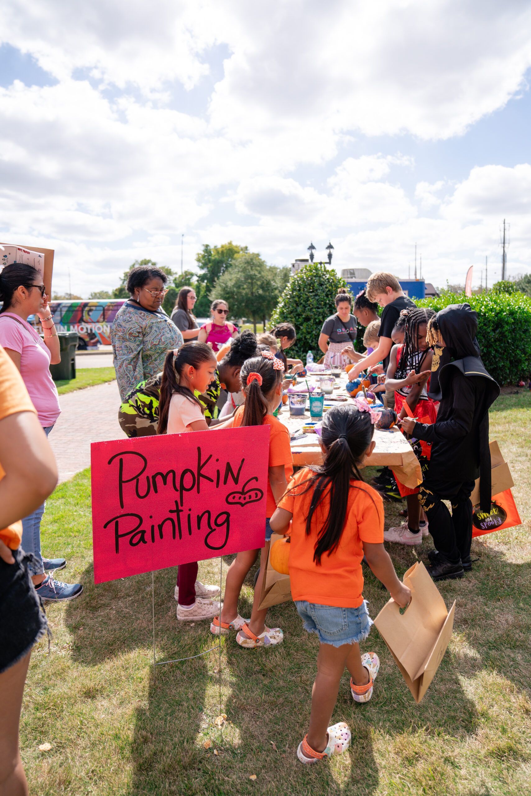 People gathered outdoors at a pumpkin painting booth with a pink sign and pumpkins on a grassy field.