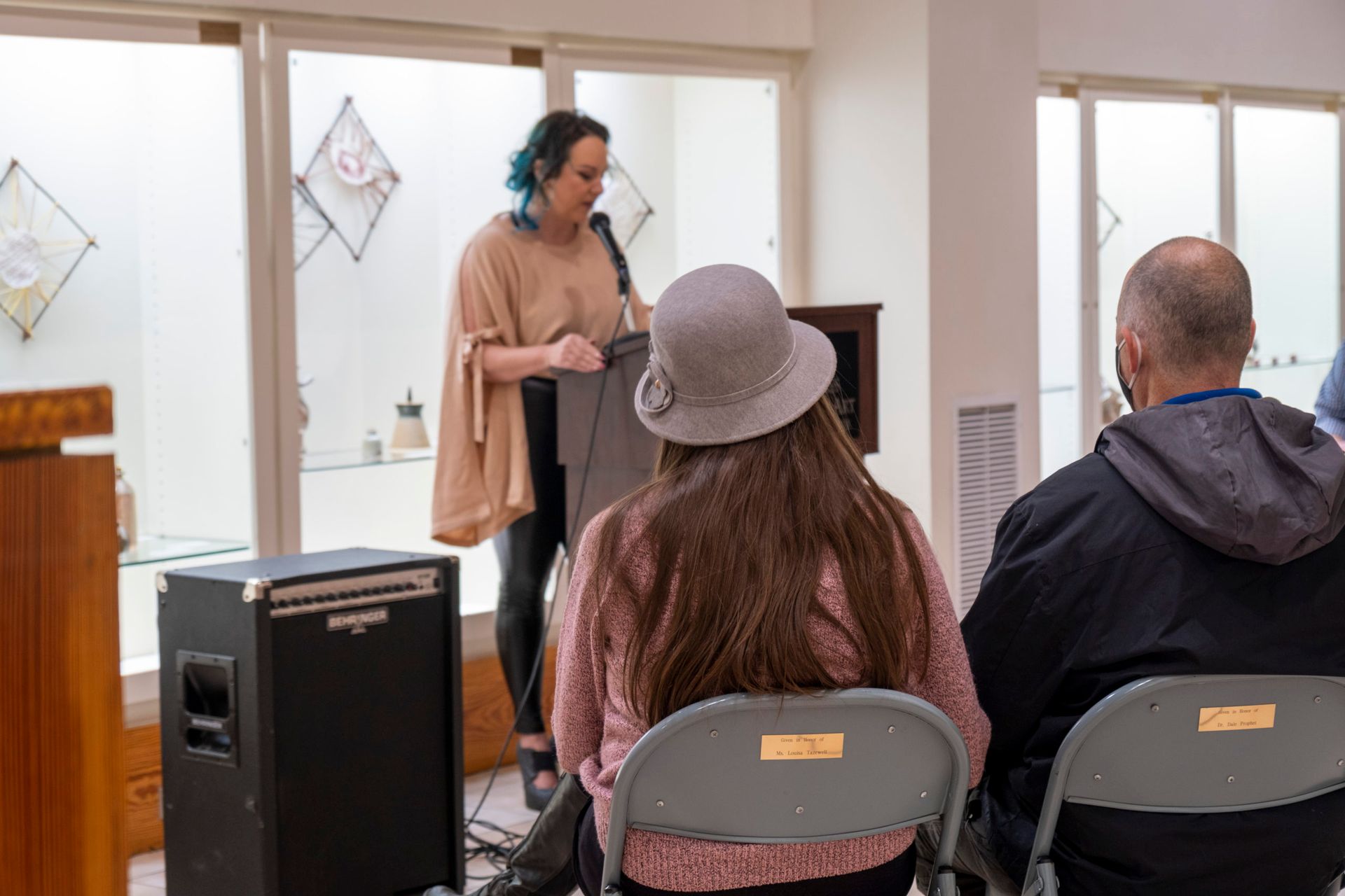 Speaker addressing seated audience in a bright room, with windows and a black speaker cabinet nearby