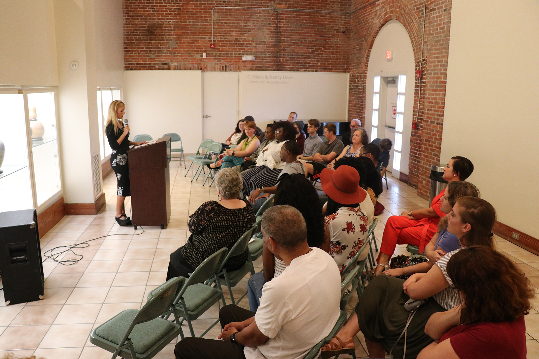 Audience seated in a brick-walled room, listening to a speaker at a podium near a window.