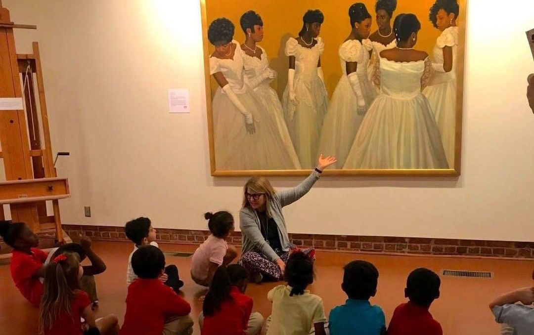 Children sitting on the floor in a museum, raising hands toward a large painting of dancers