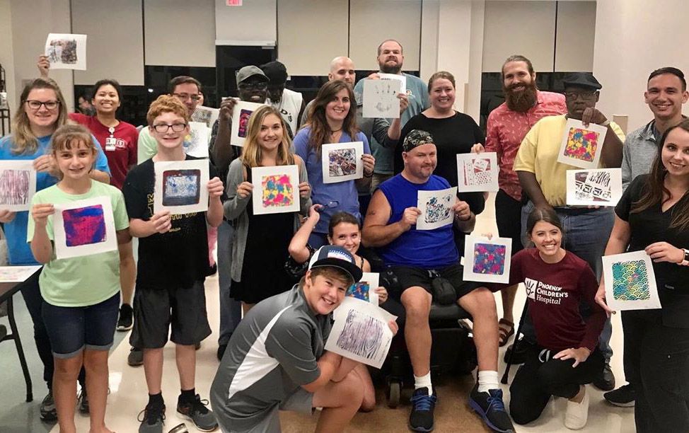 Group of people in a gym holding colorful artwork, posing together and smiling.