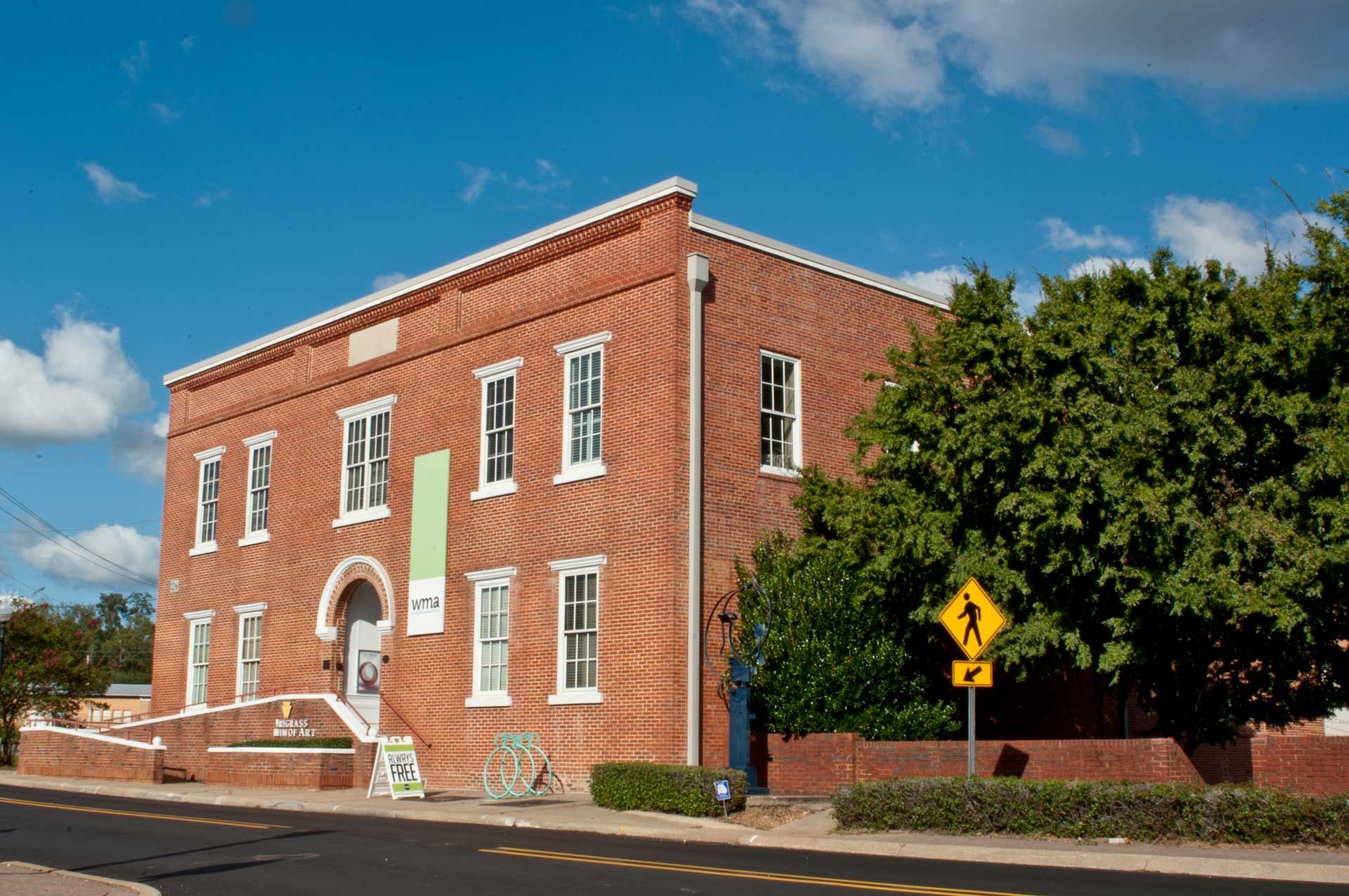Red brick building on a street corner beside a yellow road sign and large tree under a blue sky