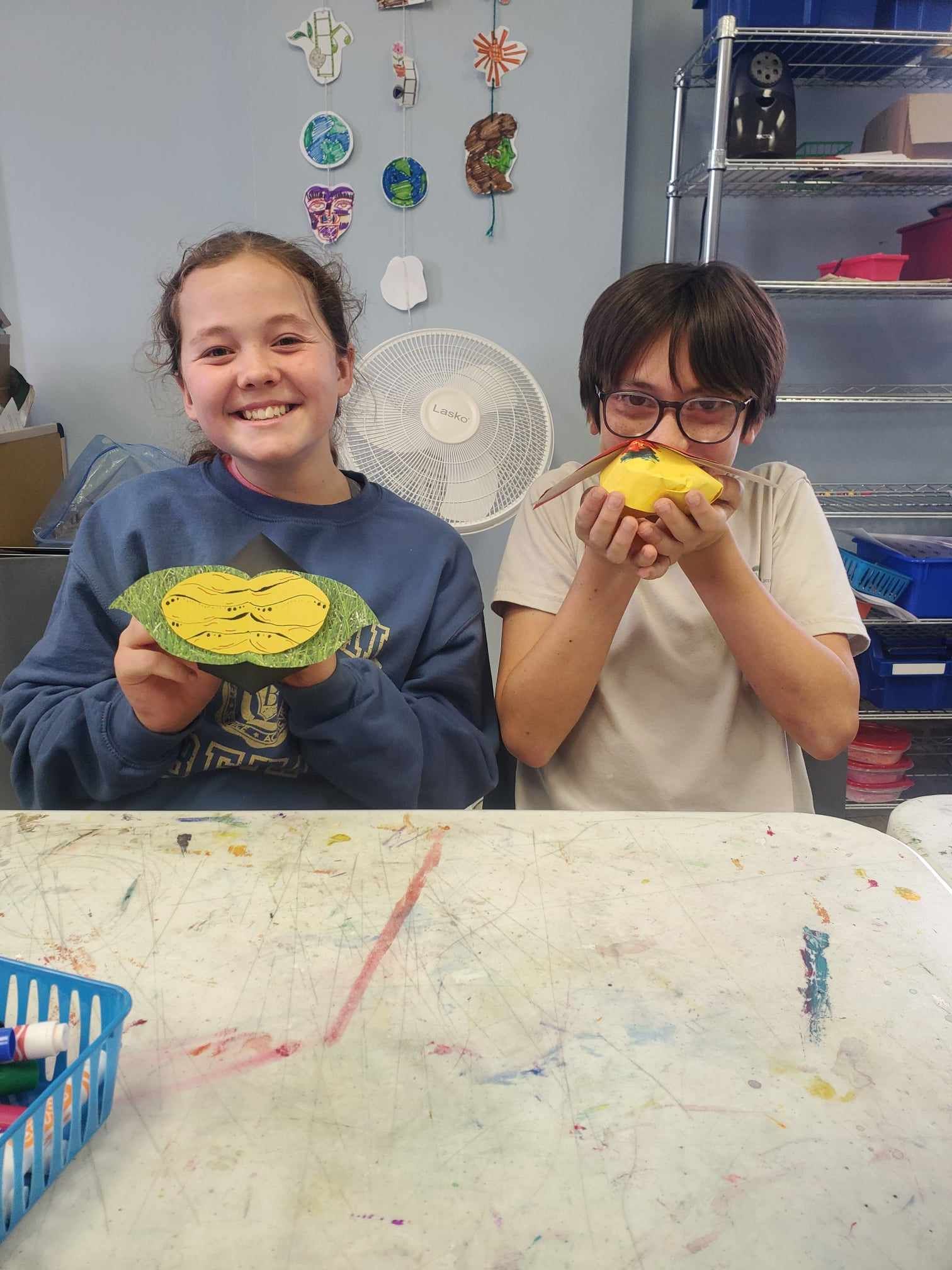 Two children smiling and holding colorful craft bowls at a classroom table