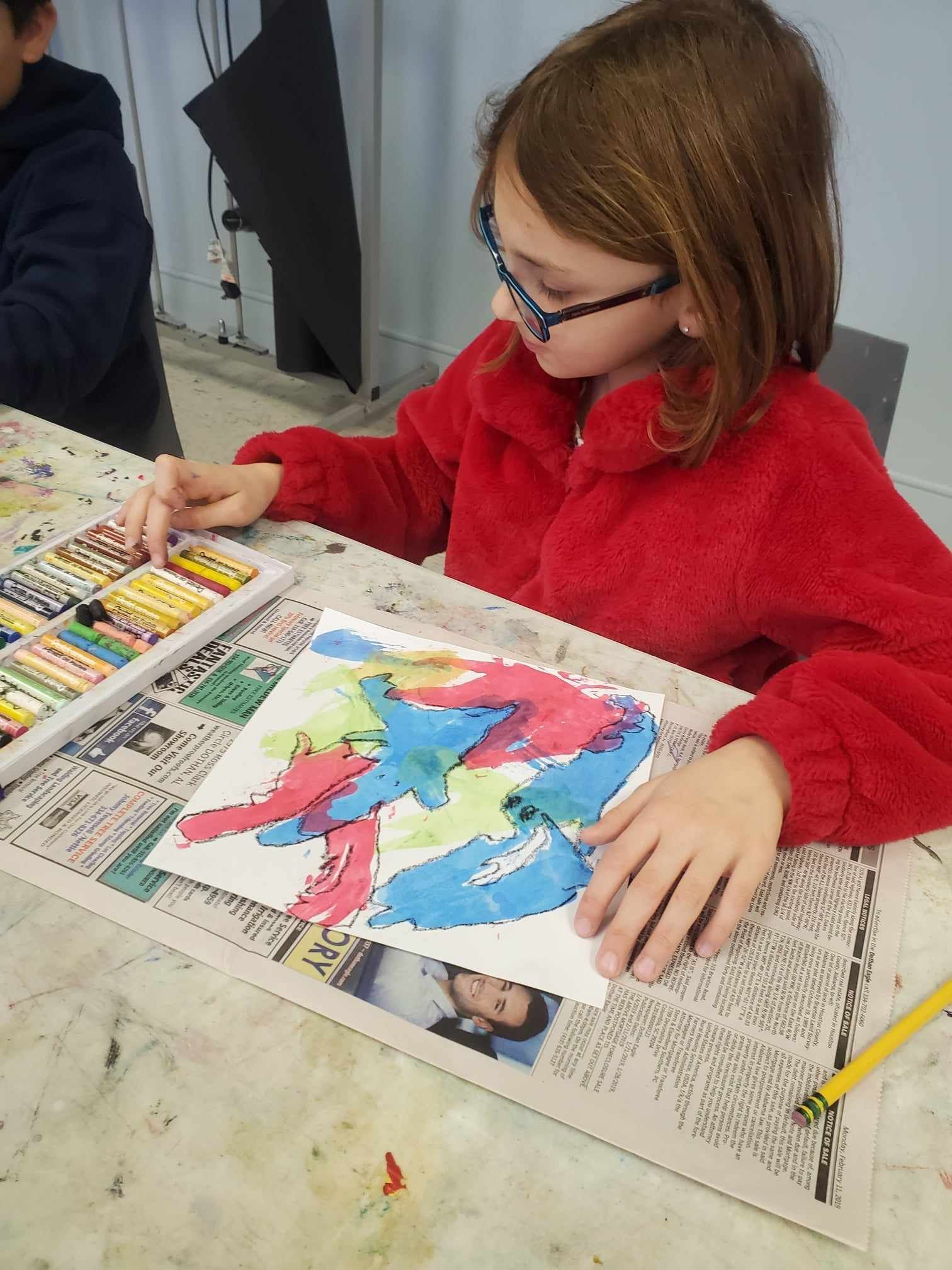 Child in red sweater painting a colorful artwork at a table with paints and newspaper spread out.