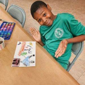 Child smiling and waving at a table with a drawing and paint set in a classroom