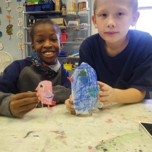 Two children holding colorful paper crafts at a table in a classroom, smiling at the camera