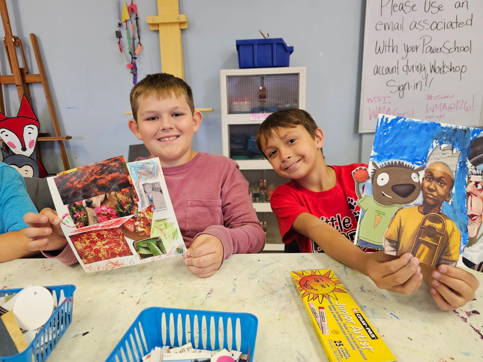 Two children smiling at a classroom table, each holding a colorful art book.