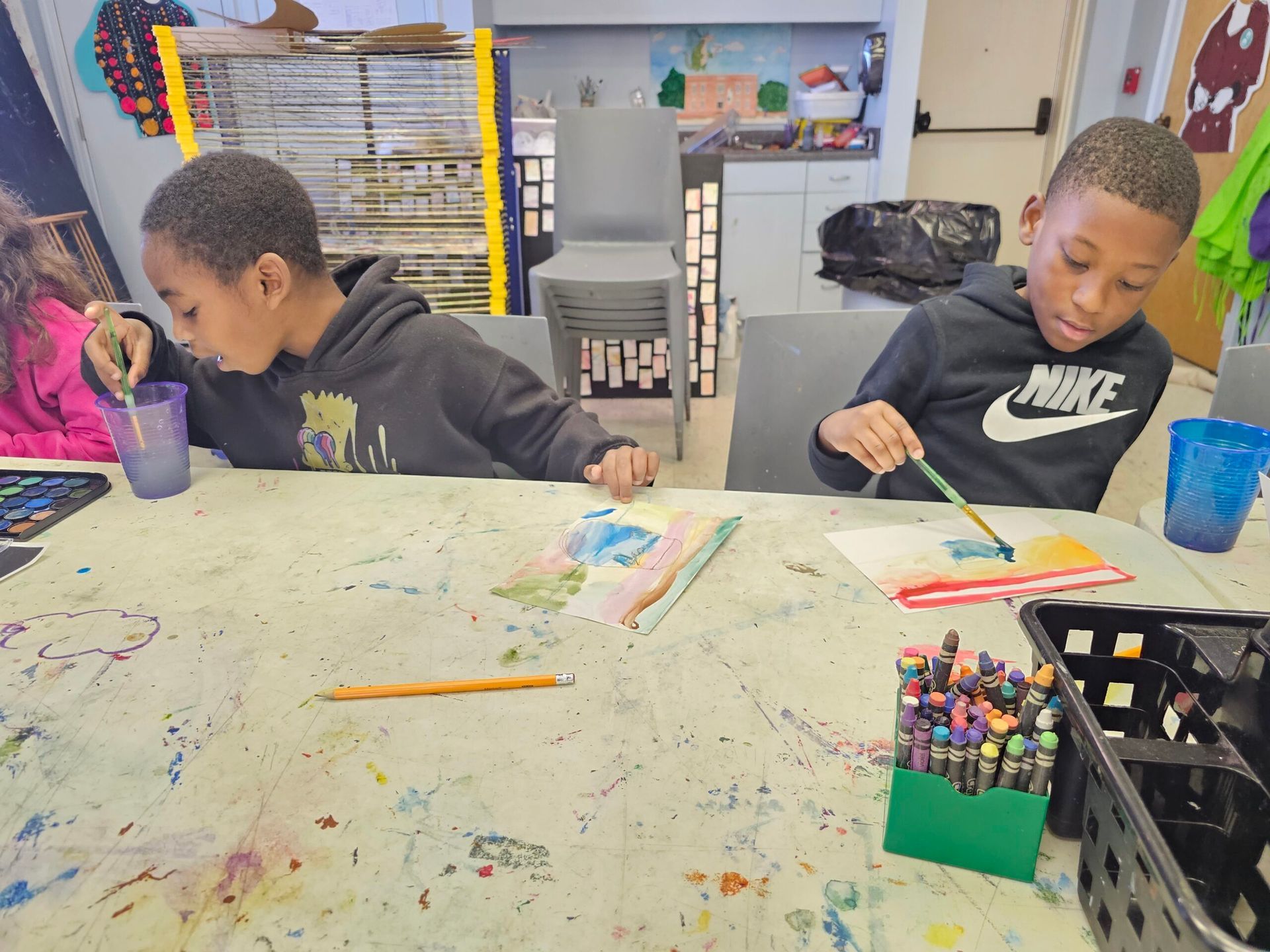 Children painting at a messy art table in a classroom, with crayons, cups, and paper.