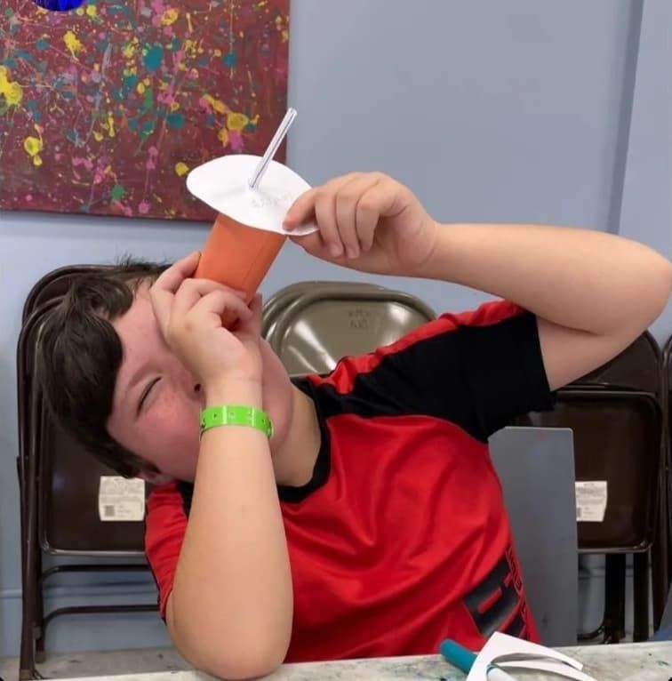 Child in a red shirt sipping from a cup with a straw at a table, with chairs and a colorful wall behind.