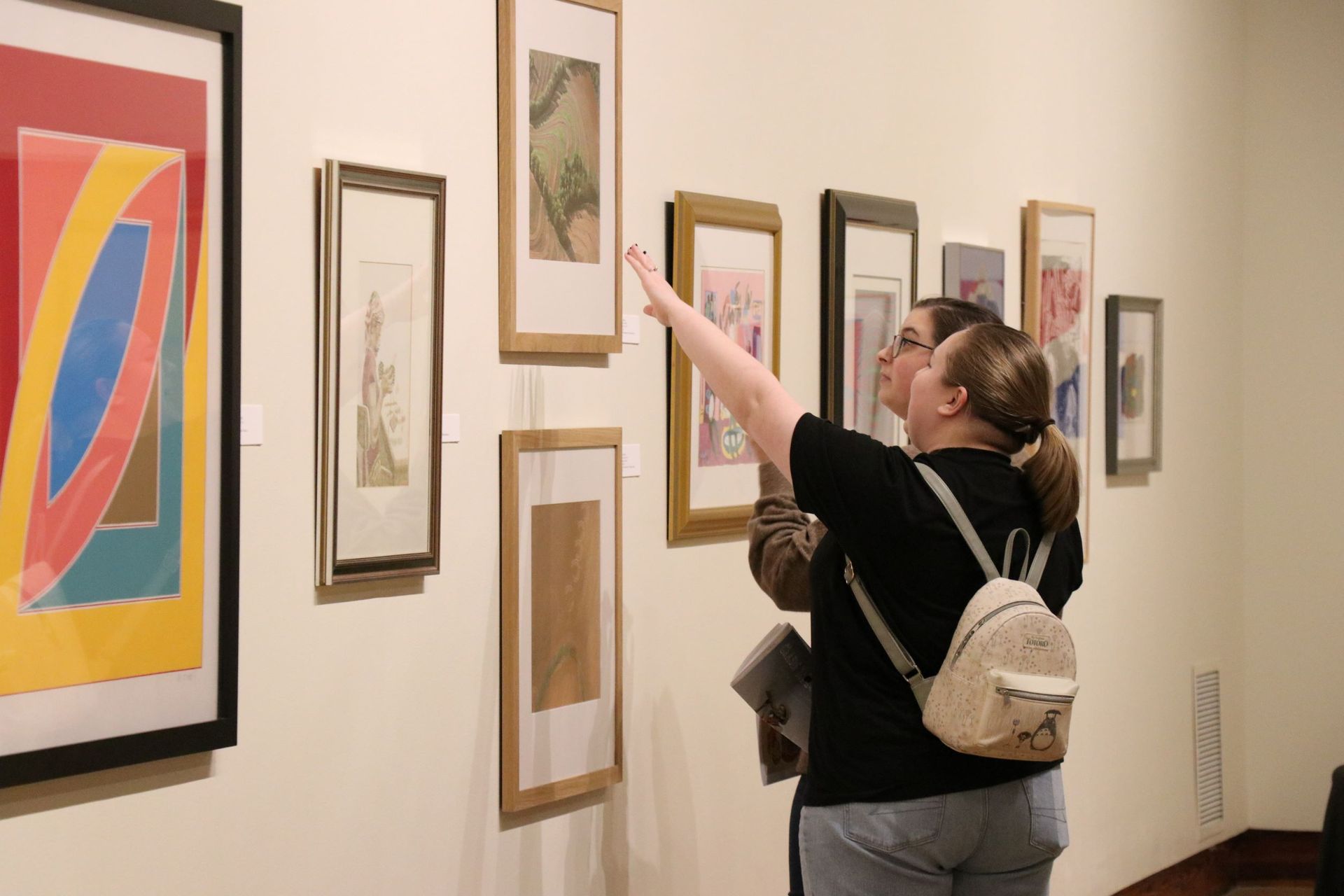 Two people viewing framed art in a gallery, one pointing at a piece on the wall.