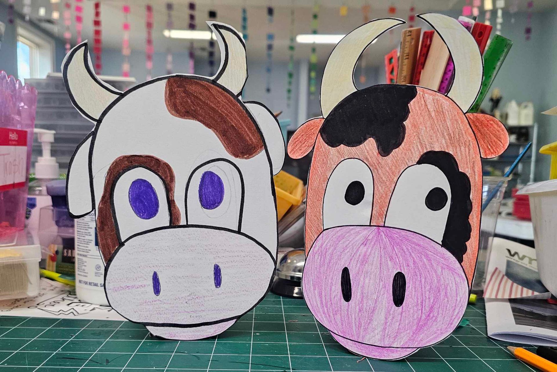 Two handmade cartoon cow masks, one white-brown and one black-pink, on a craft table