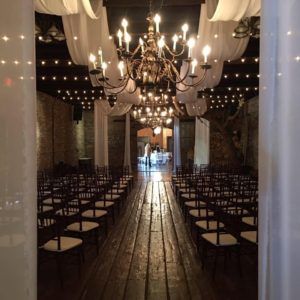 Elegant indoor event hall with chandeliers, draped ceiling, string lights, and rows of chairs along a wooden aisle