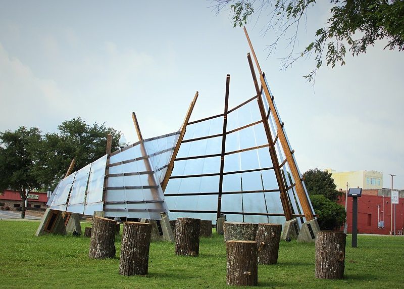 Outdoor sculpture with a curved wooden frame and metal panels on a grassy lawn, supported by stump-like bases