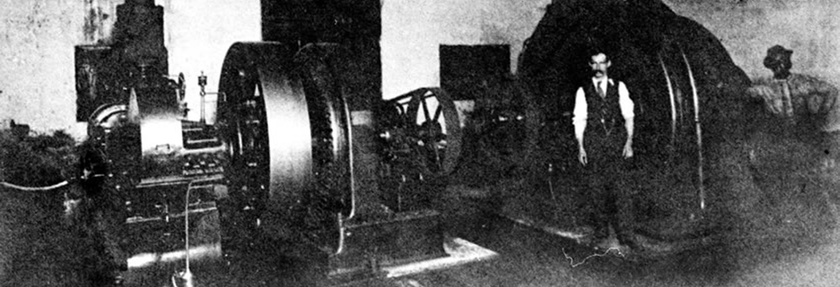 Black-and-white photo of a large vintage industrial machine with a person standing beside it in a factory-like room
