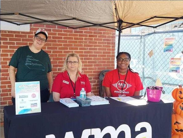 Three people at a booth under a canopy, with a black tablecloth and promotional materials.