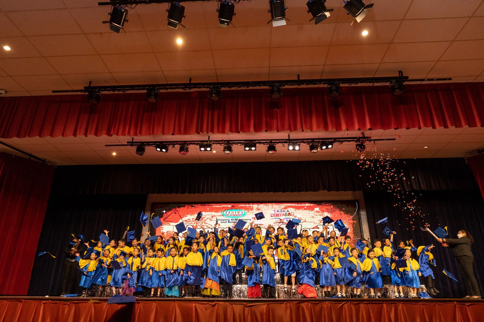 Children in blue and yellow graduation attire on a stage, throwing caps into the air. Red curtains and stage lighting.
