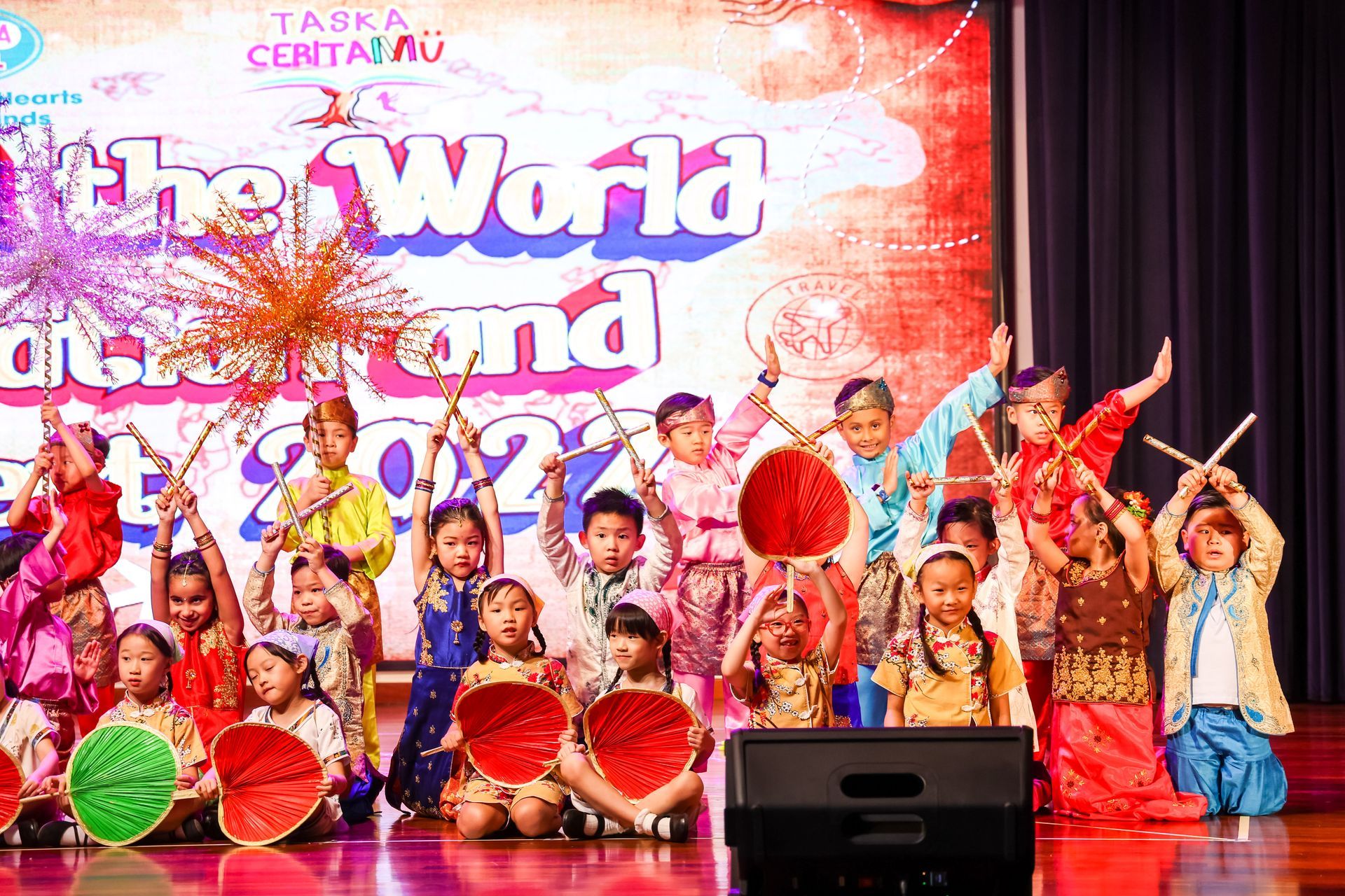 Children in colorful costumes perform on stage, holding props during a school event.