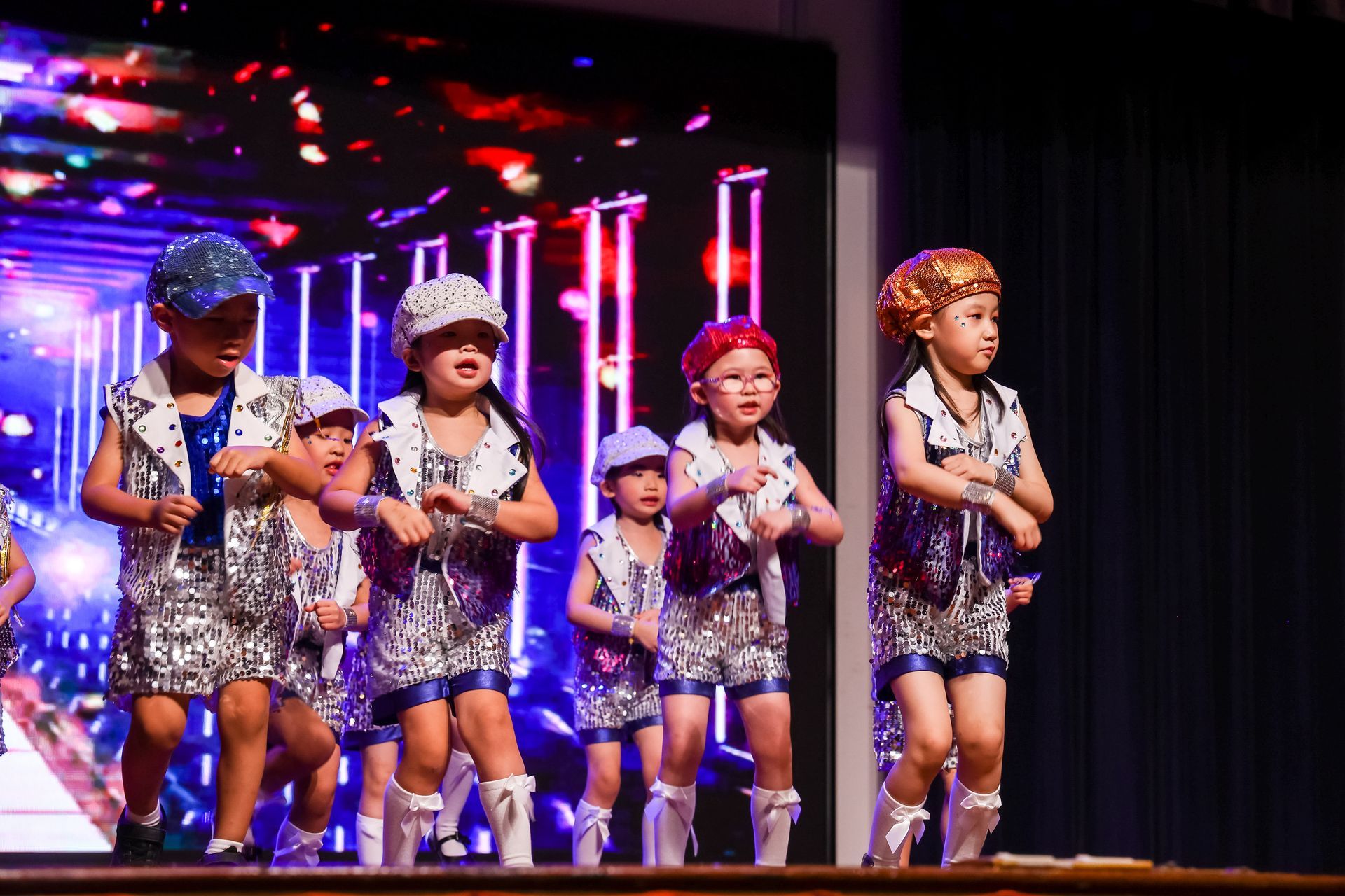 Children in matching silver outfits dance on a stage with a screen displaying colorful lights.
