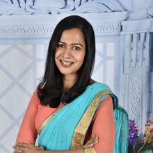 Woman in coral top and turquoise sari smiling, arms crossed; against a white and blue backdrop.