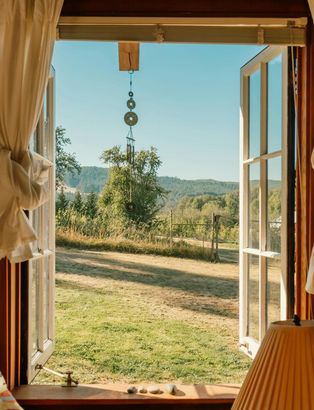 Open French doors framing a sunny countryside view with fields, trees, and hills under a blue sky.