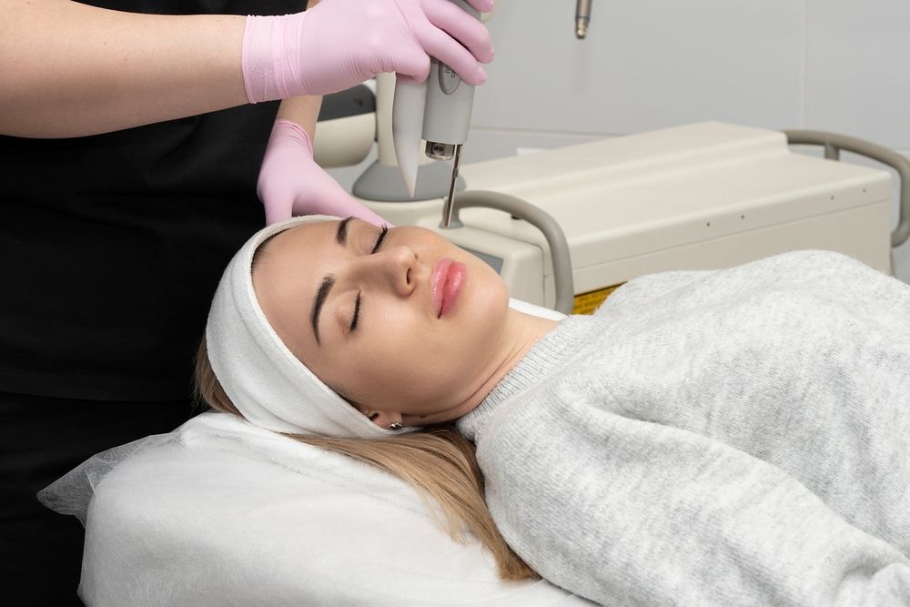 Woman receiving facial treatment; a technician with pink gloves holds a device to her face in a medical setting.