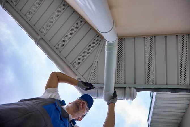 Technician cleaning a gutter on a house roof, wearing gloves and a blue cap.