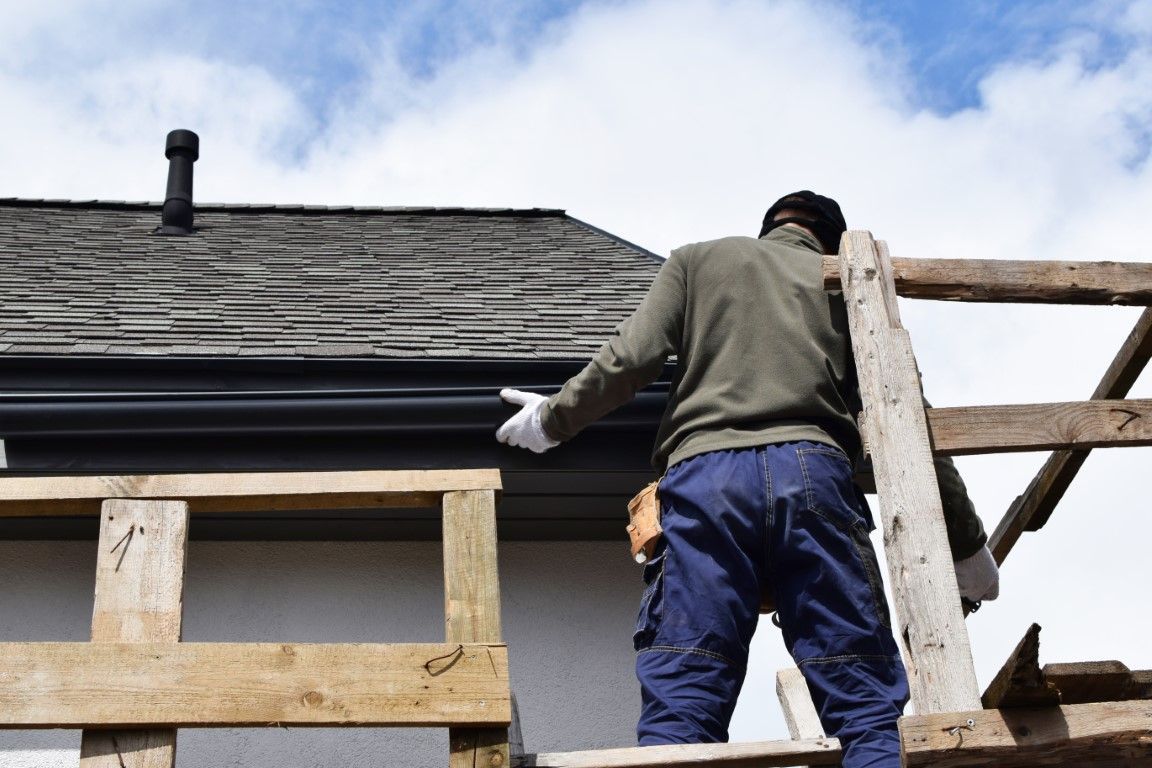Worker cleaning a roof gutter with a hose, wearing green shirt and black overalls under a blue sky.