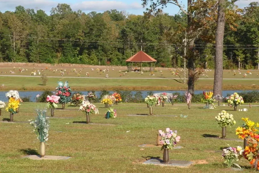 Grassy cemetery with colorful flower arrangements on graves beside a pond and trees in the background