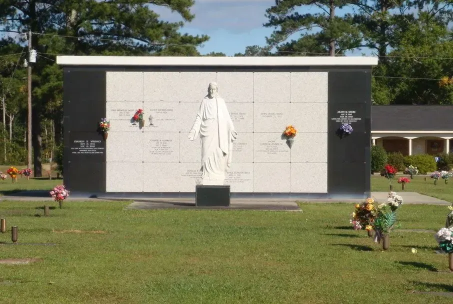 White memorial wall with a statue in front, surrounded by flowers on a grassy lawn.